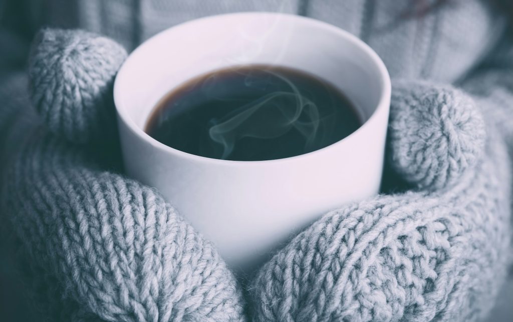 A mug being held by someone wearing mittens, representing some of the pros and cons of the winter lifestyle. By Alex Padurariu.
